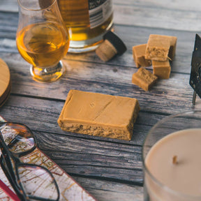 An Ochil Malt Whisky Fudge Bar rests on a rustic wooden table beside a glass of single malt whisky, a candle, a map with glasses, and small fudge cubes, creating a warm and inviting scene.