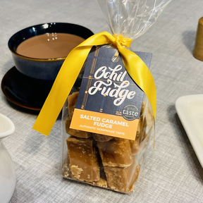 A Salted Caramel - Hand Tied Gift Bag, featuring handcrafted Scottish fudge and tied with a yellow ribbon, rests near a cup of tea or coffee and a small jug on a gray tablecloth.