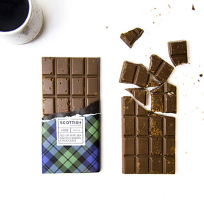 A partially unwrapped Isle of Skye Sea Salted Caramel Bar in tartan packaging sits next to a broken caramel milk chocolate bar and a cup of dark beverage, displayed on a white surface.