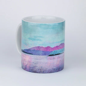 The Arran Over Sound of Bute Mug by Cath Waters features a pastel landscape of Arran with mountains, water, and a cloudy sky in blue, pink, and purple tones. The ceramic mug is shown against a plain white background.