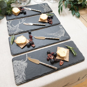 The 4 Mini Slate Cheese Board & Knife Sets - Country Animals feature rectangular boards with animal engravings, each elegantly displaying crackers, butter, berries, nuts, and herbs on a light tablecloth.