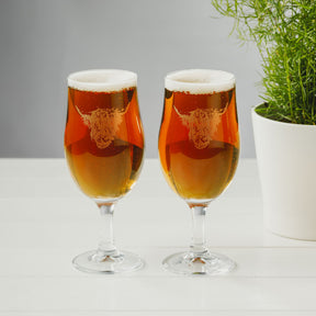 Two glasses from the Highland Cow Craft Beer Glasses (Set of 2), each featuring an etched highland cow design, sit with foamy amber beer on a white surface next to a potted green plant—making them an ideal gift for any craft beer enthusiast.