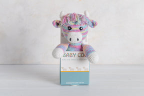 A pastel crocheted Baby Coo Crochet Coo plush with a multicolored mane sits before a white "BABY COW" box on light wood, making a charming Scottish gift.