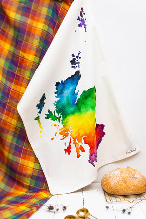 The Scotland Watercolour Map Tea Towel, featuring a vibrant rainbow design, hangs beside tartan fabric. A round loaf of bread and gold spoons sit nearby—making it the perfect unique Scottish gift.
