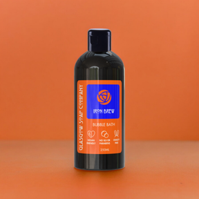 A black bottle of Iron Brew Bubble Bath sits on an orange surface and background. Featuring orange and blue labeling, this vegan bath product is infused with Irn Bru fragrance—an ideal Scottish gift by Glasgow Soap Company.