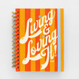 Spiral-bound 12 Month Medium Planner - Living and Loving It with orange and yellow vertical stripes, featuring the text “Living & Loving It!” and “2027” in white script.