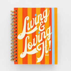 Spiral-bound 12 Month Medium Planner - Living and Loving It with orange and yellow vertical stripes, featuring the text “Living & Loving It!” and “2027” in white script.