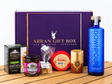 A Deluxe Scottish Gin Lover Gift Box – 70cl on a wooden surface containing a bottle of Downpour Scottish Dry Gin, a box of oatcakes, a jar of chutney, a bag of Scottish tablet, and two rounds of Arran cheddar.
