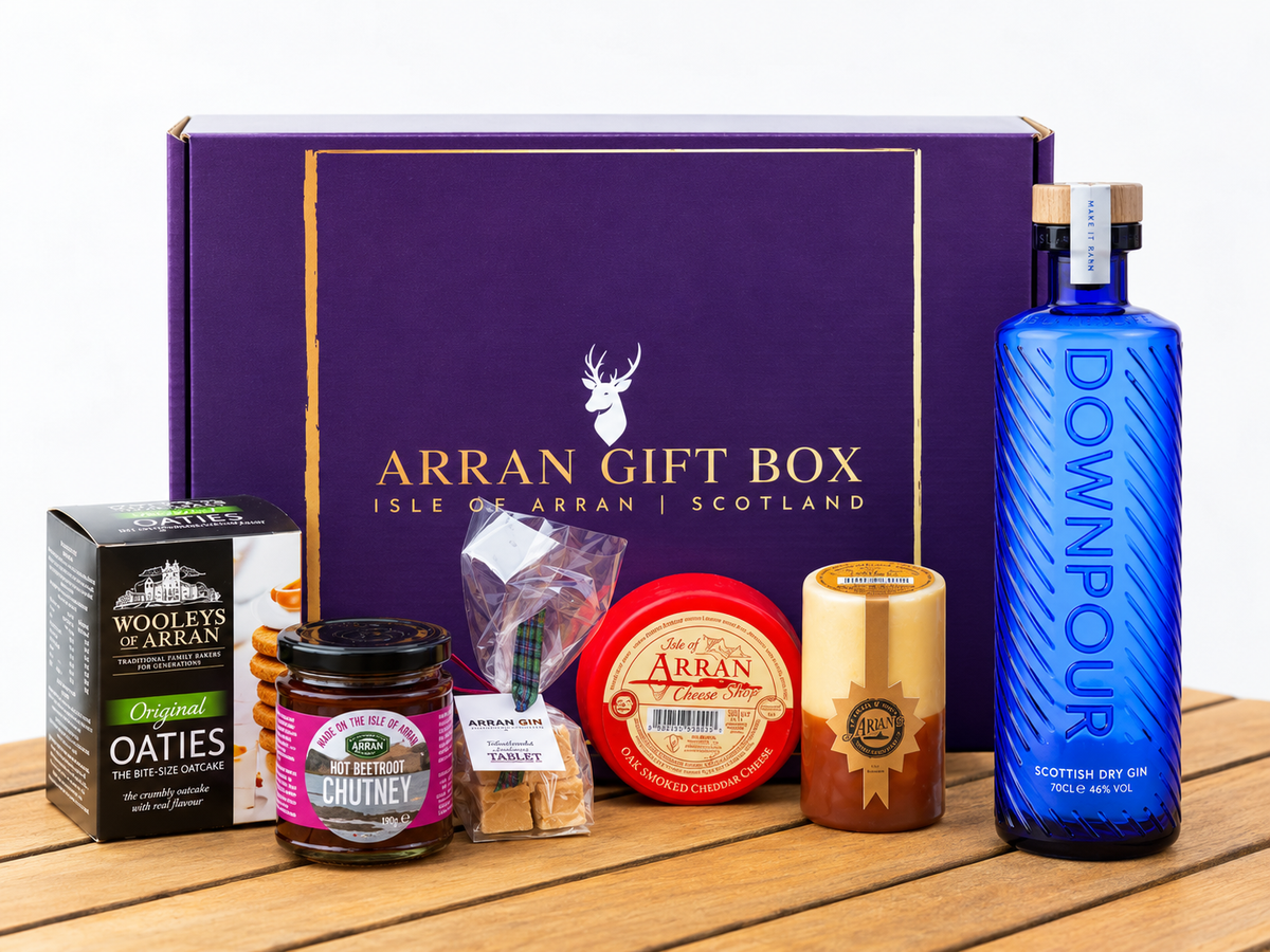 A Deluxe Scottish Gin Lover Gift Box – 70cl on a wooden surface containing a bottle of Downpour Scottish Dry Gin, a box of oatcakes, a jar of chutney, a bag of Scottish tablet, and two rounds of Arran cheddar.