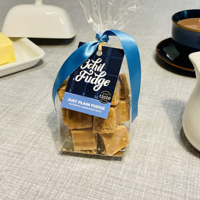 A clear hand-tied gift bag labeled "Just Plain Fudge" holds crumbly Scottish fudge cubes and rests on a gray tablecloth near butter, coffee, and a white pitcher.