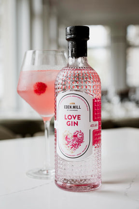 A bottle of Eden Mill Love Gin 70cl sits on a white surface next to a glass of pink gin cocktail with a raspberry garnish, set against a softly blurred, bright modern interior.