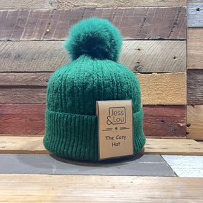 A Green Unisex Cable Bobble Hat with a pom-pom rests on a wooden surface. A brown tag attached to the hat reads “Jess & Lou The Cosy Hat.” The background features multicolored wooden planks.