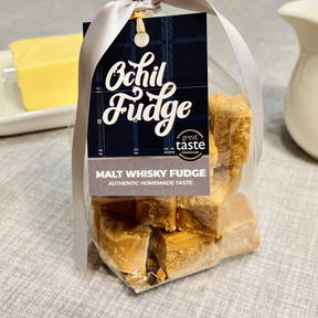A clear "Malt Whisky Fudge - Hand Tied Gift Bag" by Ochil, featuring handmade fudge cubes, a dark plaid tag, and white ribbon. Butter and a white jug sit behind on a light gray surface.