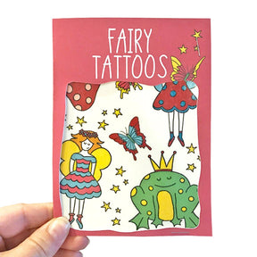 A hand holds a Fairy Transfer Tattoos package showing colorful fairies with wings, a crowned frog, butterflies, stars, and mushrooms—ideal fairy transfer tattoos for party bag surprises.