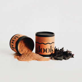 A cylindrical container labeled "dook" lies on its side, spilling Soothing Red Clay Mask powder next to an upright matching container and dark seaweed, all on a white surface—ideal for sensitive skin.