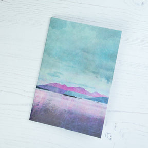 The Arran over Sound of Bute A6 notepad features a watercolor landscape of purple mountains, a reflective lake, and a blue-green sky—printed by a UK company and shown on a white wooden surface.