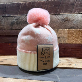 A Unisex Star Cosy Bobble Hat in pink and white, featuring a fluffy pom-pom on top, rests on a wooden surface. The hat has a tag that reads “Jess & Lou - The Cosy Hat,” with wooden planks in the background.