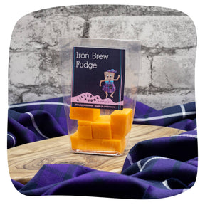 A clear bag of Iron Brew Scottish Fudge filled with orange fudge pieces rests on a wooden board, set atop purple tartan fabric with a gray brick wall as the backdrop.