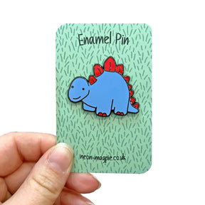 A hand holds a card with a Dinosaur Enamel Pin Badge, featuring a blue and red dino. The grass-green card reads "Enamel Pin" at the top and "neon-magpie.co.uk" at the bottom, highlighting this playful Neon Magpie accessory.