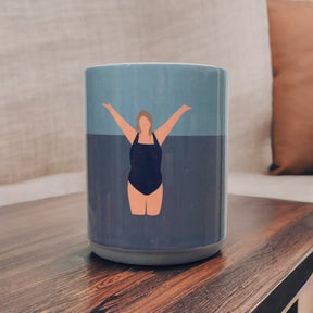 The I Love the Water Mug features a graceful woman in a dark swimsuit, arms raised against a blue background. Displayed on wood with a beige couch and brown pillow behind. Dishwasher safe for easy cleaning.
