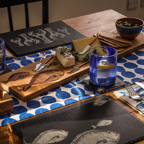 A wooden dining table set with blue and white polka dot placemats, slate fish-design plates, an Oak Large Platter - Fish Repeat for cheese and crackers, cutlery, and lemon water—an ideal wedding gift.
