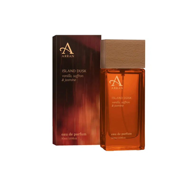 A bottle of Island Dusk Eau de Parfum 50ml sits next to its sleek, dark gradient box with gold text. This luxury fragrance by Arran Sense of Scotland features notes of vanilla, saffron, and jasmine and has a stylish wooden cap.