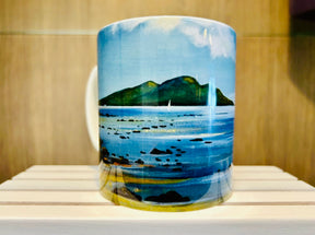 The Lamlash to Holy Isle Mug showcases an illustrated seascape with calm blue water, scattered rocks, a distant green island, and a white sailboat beneath a partly cloudy sky, all set against a wooden background.