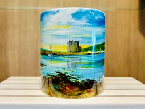 The Lochranza Castle Mug showcases a vibrant painted landscape of a castle by a lake with a sailboat, green hills, and a bright sky, all set against a white wooden surface.