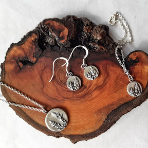 The Machrie Heather Earrings, made of sterling silver and featuring a round pendant with a raised botanical design, are displayed on a polished brown wooden slab set against a white background.