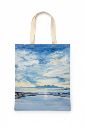 The Reflections of Arran Tote Bag has beige handles and displays a painted seascape with a cloudy sky, tranquil blue water, and distant mountains on the horizon.
