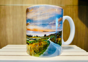 The Road to Kildonan Mug is a white mug featuring a vibrant landscape painting of a winding road leading to water at sunset, surrounded by trees and greenery under a colorful sky.