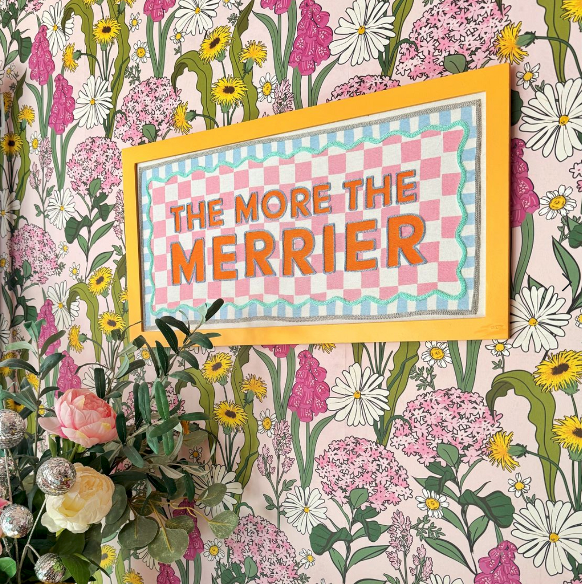 The More the Merrier Orange Framed Fabric Sign featuring orange text on a pink and white chequered background, mounted on a floral wall.