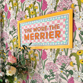The More the Merrier Orange Framed Fabric Sign featuring orange text on a pink and white chequered background, mounted on a floral wall.