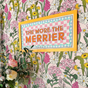 The More the Merrier Orange Framed Fabric Sign featuring orange text on a pink and white chequered background, mounted on a floral wall.