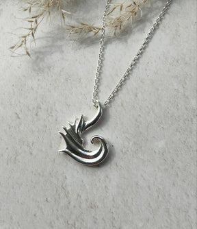 The "Tide Keeper" Necklace is a sterling silver pendant featuring a stylized dragon or wave, shown on a light textured surface with dried grasses in the background.