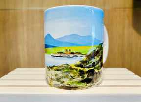 The Waverley Memories Mug is a ceramic mug featuring a painted landscape of a boat on a lake, green hills, and blue mountains under a clear sky, displayed on a white wooden surface.
