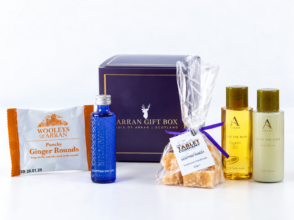 A Wee Goatfell Scottish Gin Gift Box 5cl featuring a dark blue box, a small blue bottle of gin, a bag of Scottish tablet, ginger biscuits, and Arran Sense of Scotland body wash and lotion.
