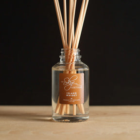 The Island Whisky Diffuser, a glass bottle with clear liquid and wooden sticks, sits on a wooden surface against a black background, capturing the unique Scottish scent by Isle of Skye Candle Company.