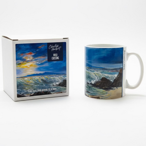 A "Wild Crossing Mug" and its matching box showcase ocean waves crashing against rocks beneath a vibrant sky, with the “Wild Crossing” label displayed on both the mug and the box.