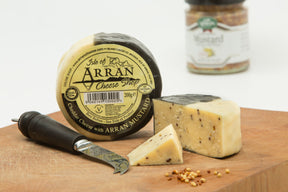 A wheel of "Arran Mustard flavoured Cheddar" with a slice that reveals wholegrain mustard seeds sits enticingly on a wooden board. Complementing this flavored cheese, a black-handled knife rests nearby, and in the background, a jar of Arran Mustard enhances the gourmet allure.