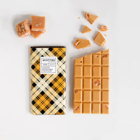A Scottish Butter Tablet Chocolate Bar, wrapped in yellow, black, and white tartan, sits on a white background beside broken chocolate pieces and chunks of tablet.