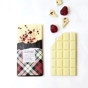 A Cranachan White Chocolate Bar with Raspberries, Whisky & Oats, partially unwrapped in a tartan wrapper, sits beside a bitten piece and fresh raspberries on a white background.