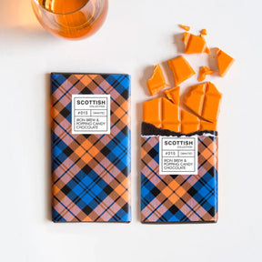 A glass of orange liquid sits beside two blue and orange tartan-wrapped Iron Brew Chocolate Bars with Popping Candy, one partially unwrapped and broken, handmade in the UK with white Belgian chocolate.